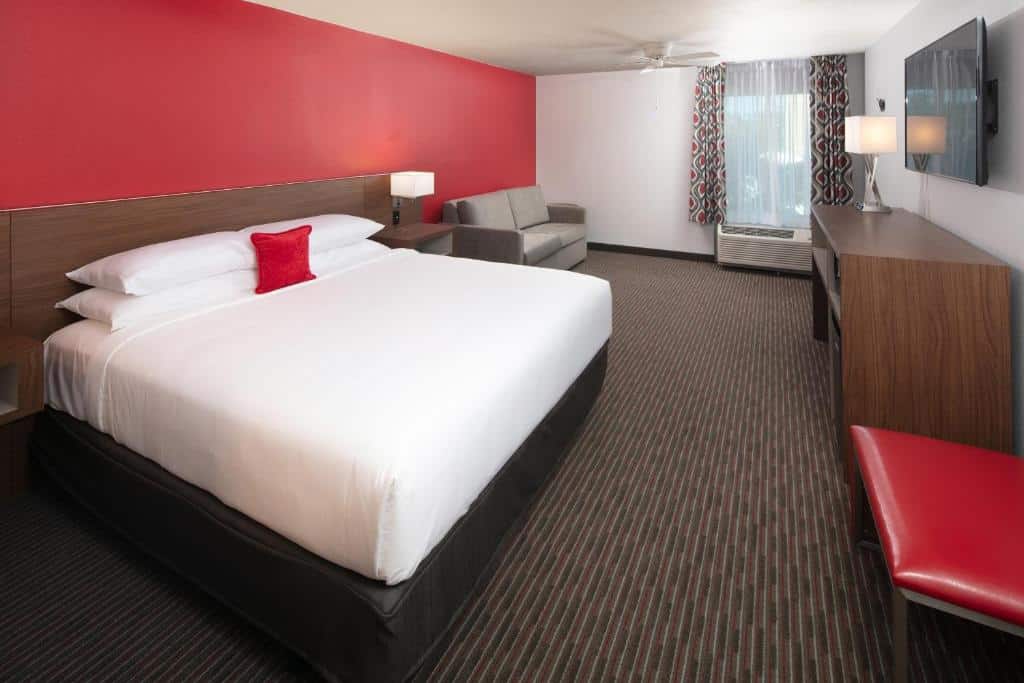 quarto amplo do Red Lion Hotel Orlando Lake Buena Vista South- Near Disney com cômoda a frente com tv com cama e mesinha e luminária de ambos os lados, com sofá e janela com aquecer abaixo