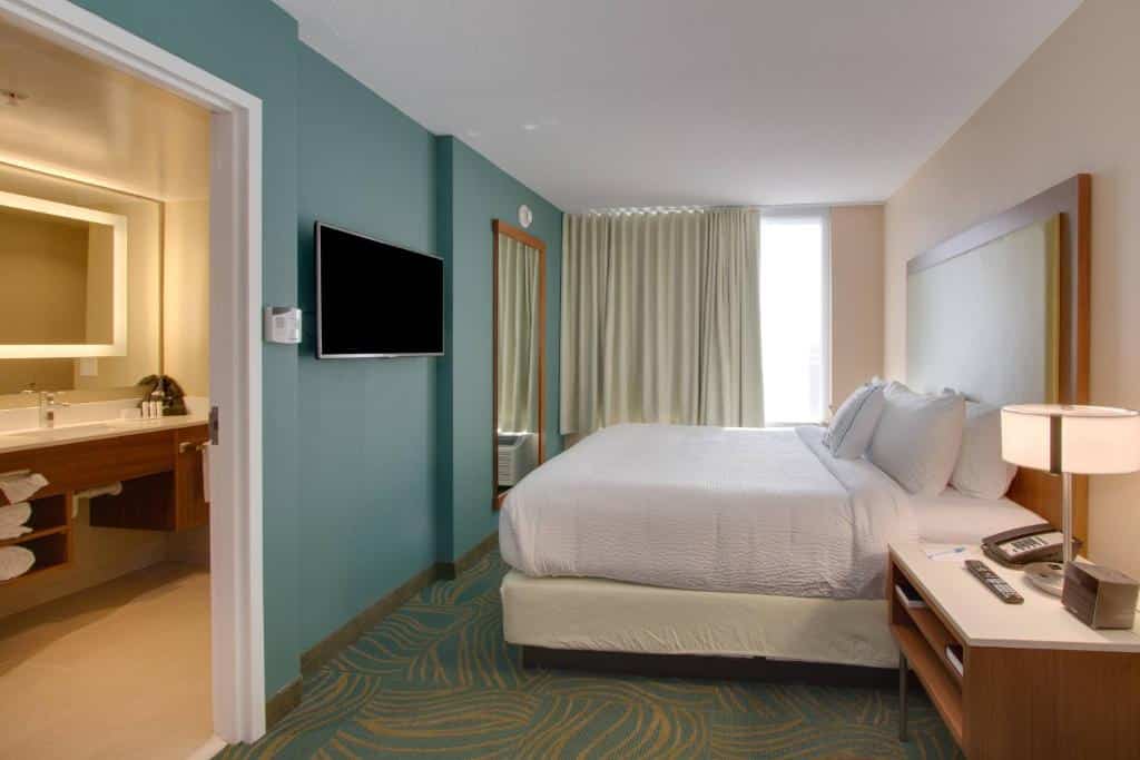 quarto do SpringHill Suites by Marriott Orlando Lake Buena Vista South com banheiro grande, tv em frente à uma cama de casal, com mesa ao lado e janela com cortinas do outro lado