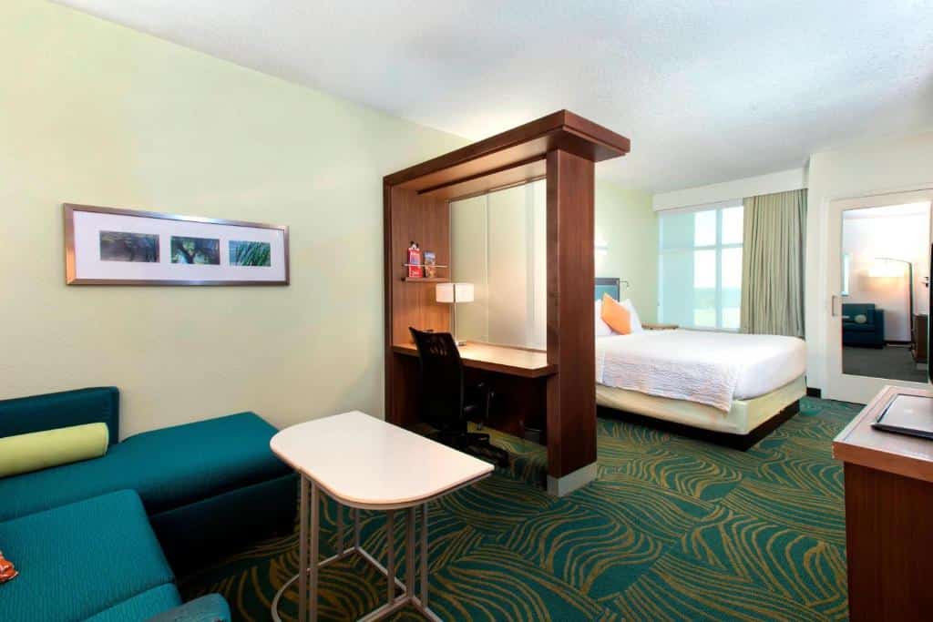 quarto do SpringHill Suites by Marriott Orlando que possui dois ambientes: uma sala de estar com sofá e mesinha em frente à uma tv e, outro com cama de casal e janela grande, separados por uma mesa com cadeira e luminária