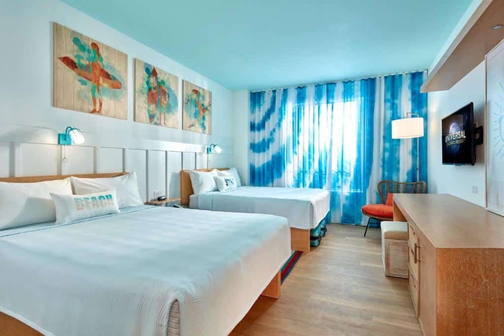 quarto amplo do Universal's Endless Summer Resort - Surfside Inn and Suites com motivos de praia e quadros de surf, duas camas com mesinha e luminária no meio delas, bancada com cadeira e tv à frente