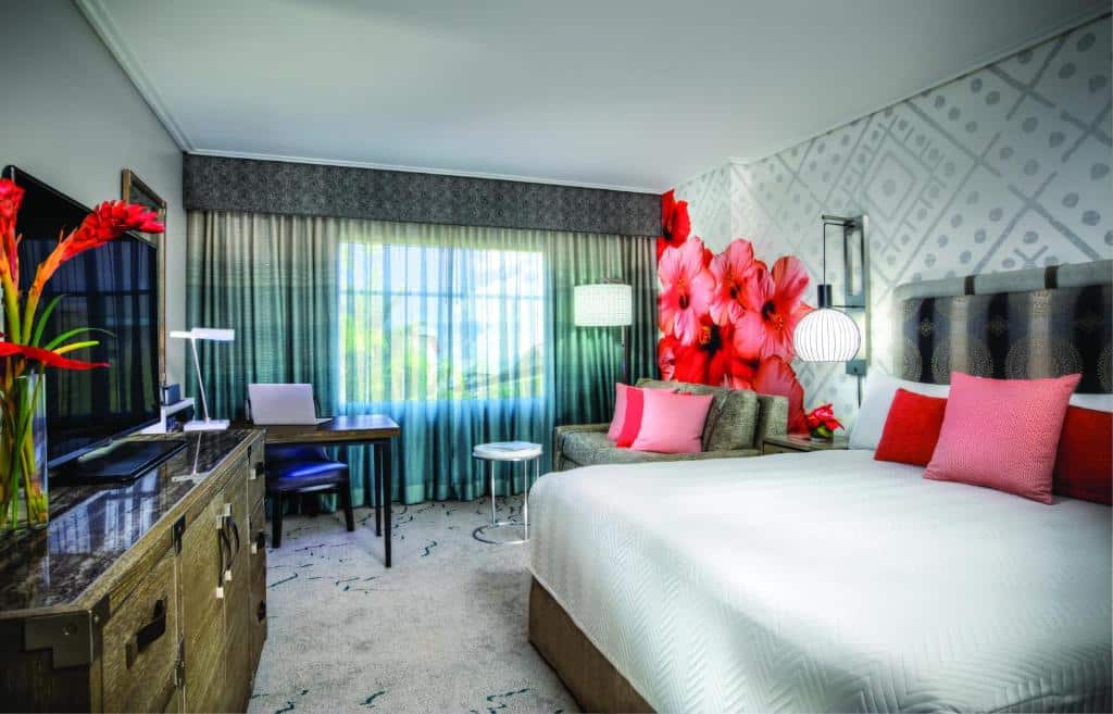 quarto do Universal's Loews Royal Pacific Resort com papel de parede de flores vermelhas, janela bem grande com cortinas com motivo de floresta, cama grande de casal com luminária e mesinha de ambos os lados, sofá ao lado e mesa com cadeiras, cômoda e tv à frente