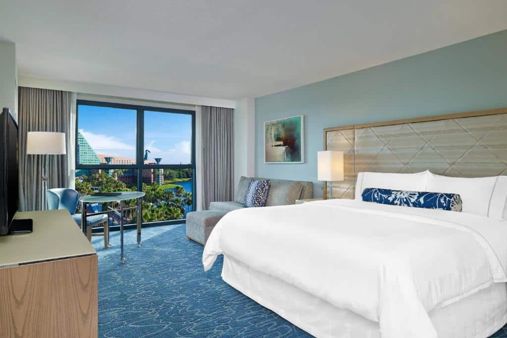 quarto amplo do Walt Disney World Swan com cama bem grande com mesinha e luminária de ambos os lados, bancada com tv à frente e uma cadeira de trabalho em frente a uma poltrona e janela bem grande com vista do lago do hotel