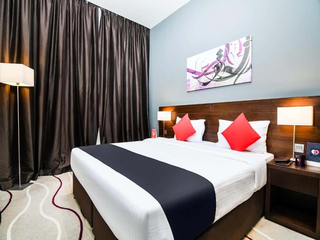 Quarto do Action Hotel Ras Al Khaimah com uma cama de casal, com mesinhas de cabeceira com abajures, um carpete bege com desenhos vinho e branco, uma janela com cortinas e um quadro sob a cama