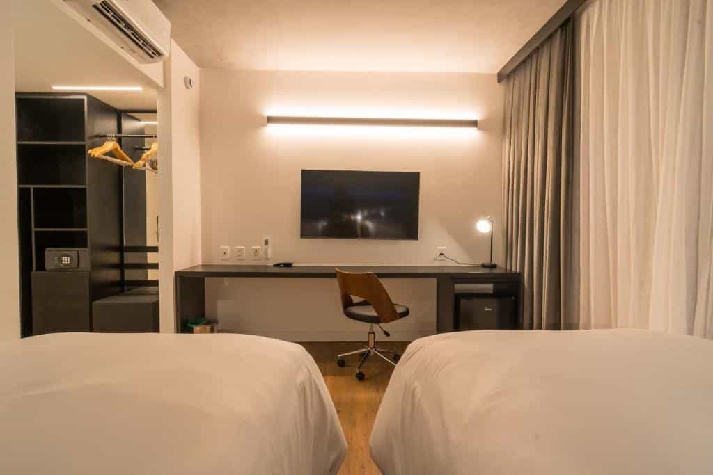 Visão de duas camas de solteiro, com uma mesa e uma cadeira na frente e uma televisão. No canto esquerdo uma parte do armário.