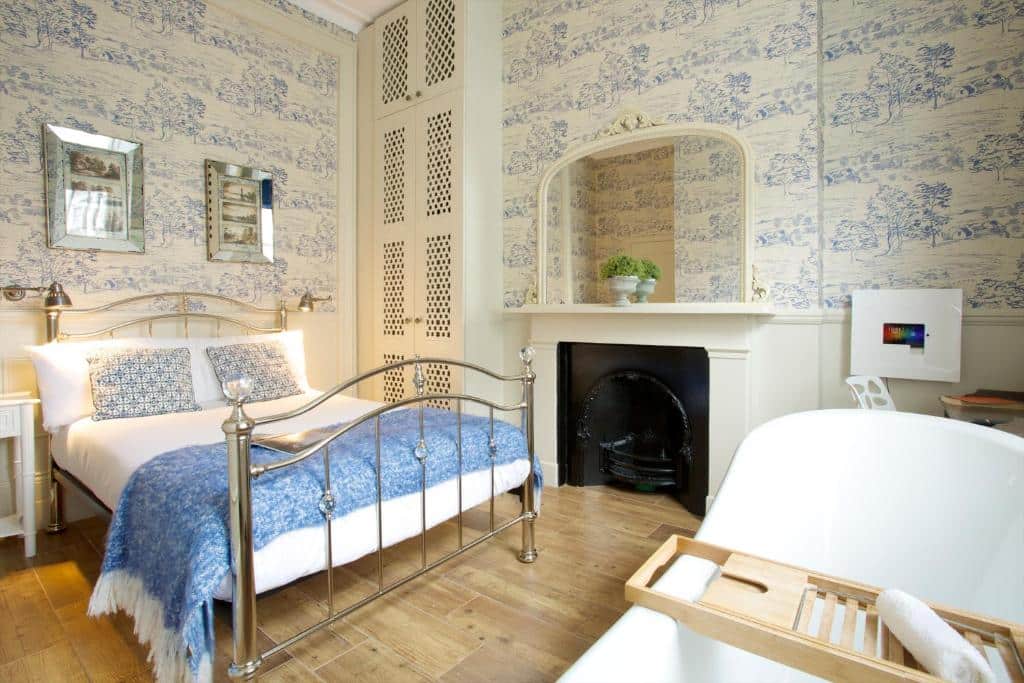 Quarto do Bower House com uma armário, uma pequena lareira, uma cama de casal em uma estrutura de ferro, uma banheira e um papel de parede branco e azul no ambiente