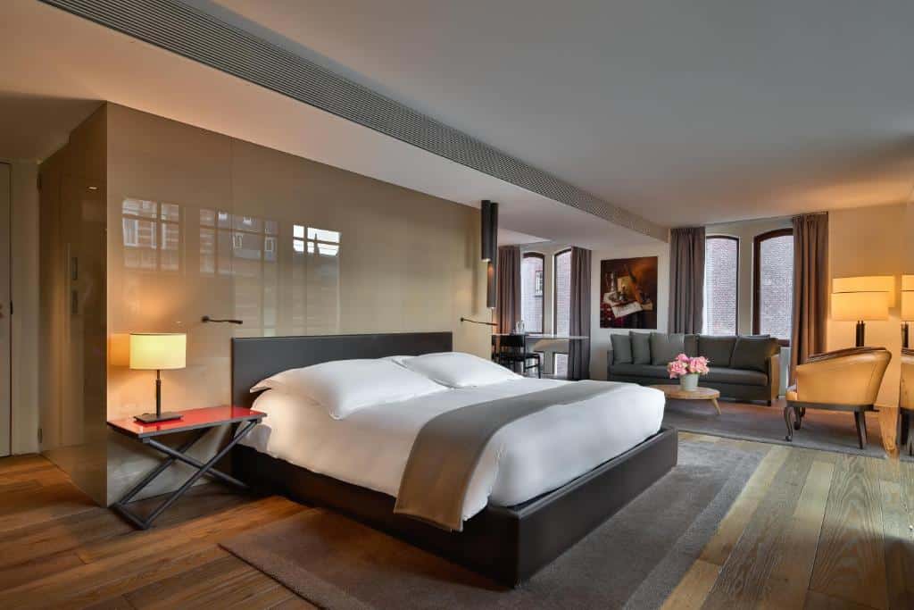 Suíte Grand Júnior do Conservatorium Hotel, de 55 m², com cama de casal extragrande, mesa com abajur ao lado da cama e, ao fundo, uma área de estar com sofás e janela com cortina