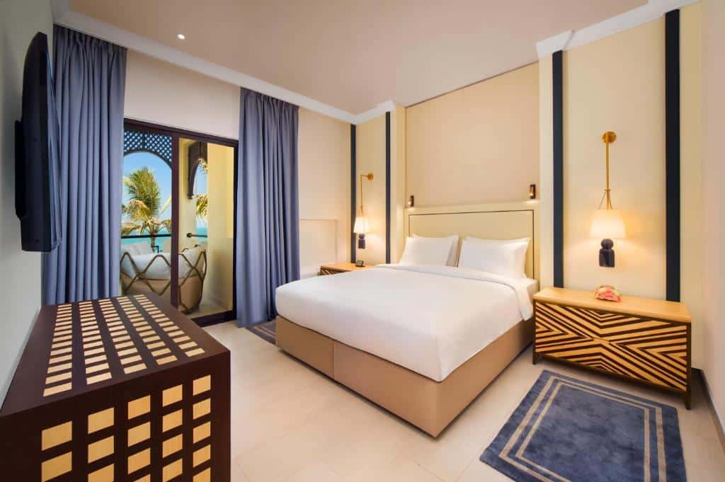 Quarto do Hilton Ras Al Khaimah Beach Resort com uma cama de casal, duas mesinhas de cabeceira com luminárias penduradas, há uma varanda com poltronas dando vista para a praia, e uma televisão de frente para a cama, para representar hotéis de luxo em Ras Al Khaimah