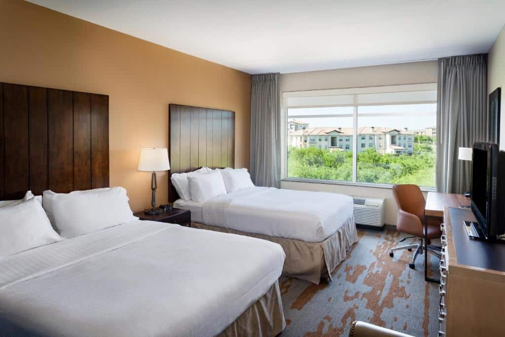 Quarto do Holiday Inn San Antonio Northwest- SeaWorld Area, an IHG Hotel com uma cama de casal e uma de solteiro, uma janela ampla com cortinas, um móvel com uma televisão em cima e uma mesa de escritório com uma cadeira giratória
