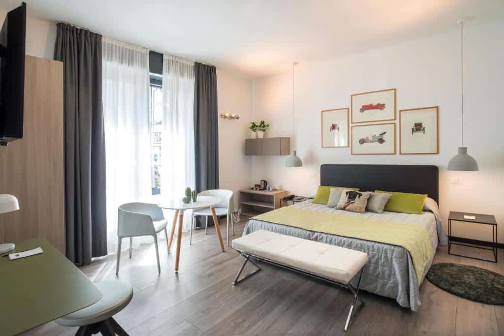Quarto espaçoso do Hotel Bernina com uma varanda com cortinas, uma cama de casal, uma pequena mesinha redonda com duas poltronas, uma mesinha de escritório com um banquinho, chão de madeira e alguns itens de decoração, como luminárias e quadros, para representar hotéis baratos em Milão
