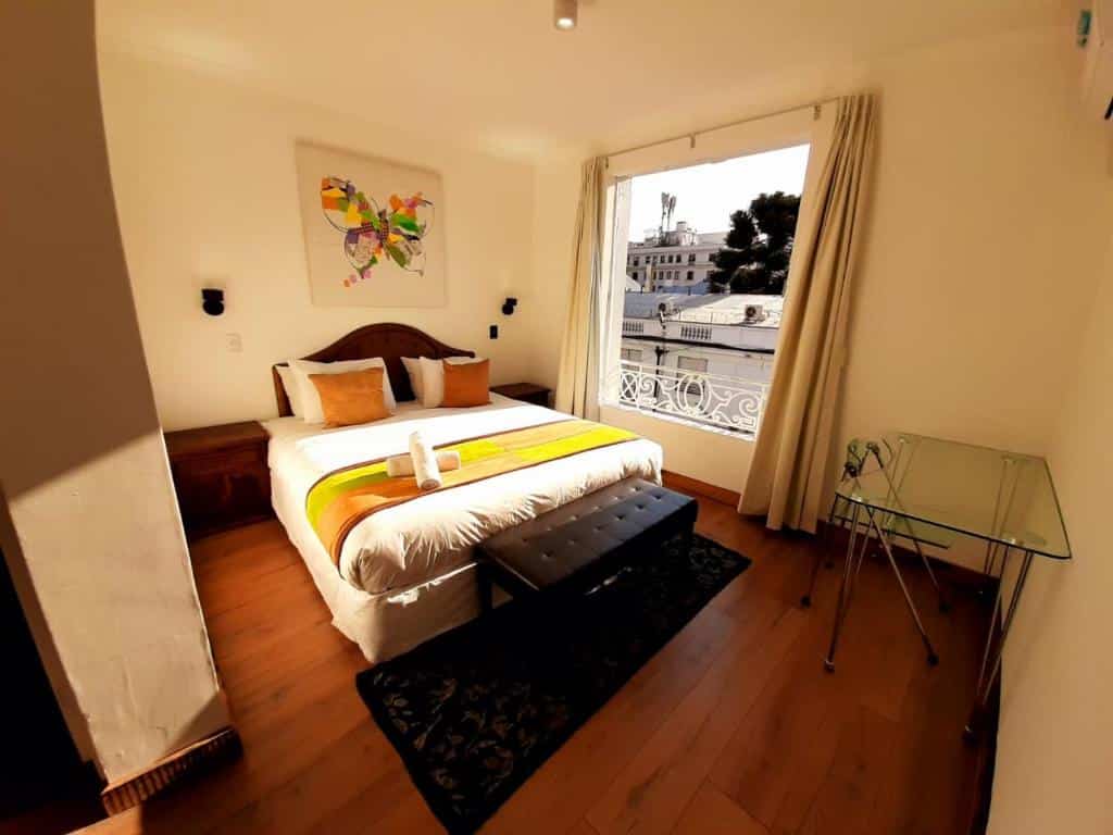 Quarto do Hotel Boutique Santiago Suite com cama de casal do lado esquerdo com duas cômodas de madeira ao lado da cama e uma mesa de trabalho de vidro em frente a cama.
