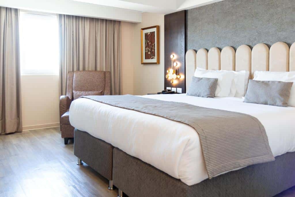 Quarto do Hotel He Wang Santiago com cama de casal a frente, do lado esquerdo da cama cômoda com telefone e uma poltrona cinza.