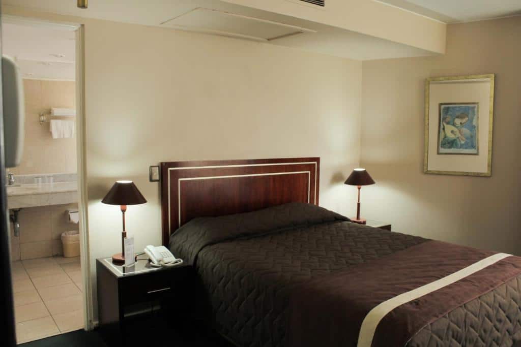 Quarto do Hotel Leonardo da Vinci com cama de casal no centro do quarto com duas cômodas de madeiras com luminárias.