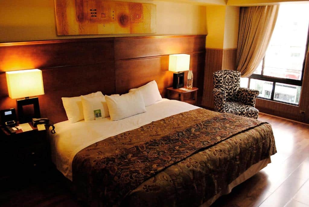 Quarto do Hotel Panamericano com cama de casal duas cômodas de madeira ao lado da cama com luminárias e uma poltrona do lado esquerdo do quarto.