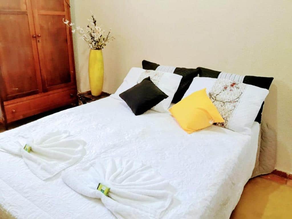 Quarto com cama de casal branca, armário de madeira e um vaso amarelo com flor.