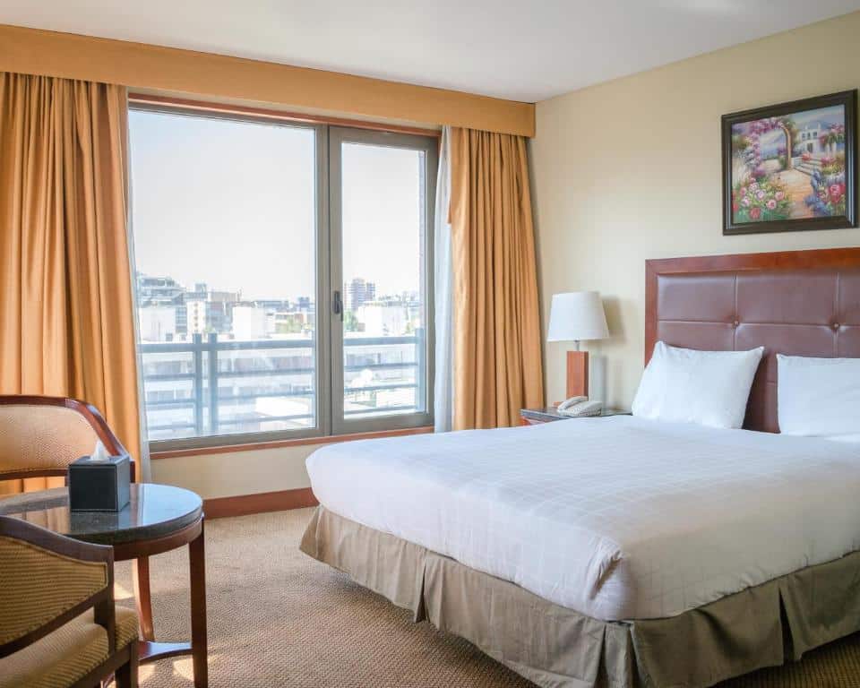 Quarto do Hotel Stanford com cama de casal a frente do lado esquerdo do quarto cômoda com luminária e telefone e em frente a cama uma mesa redonda com duas cadeiras.