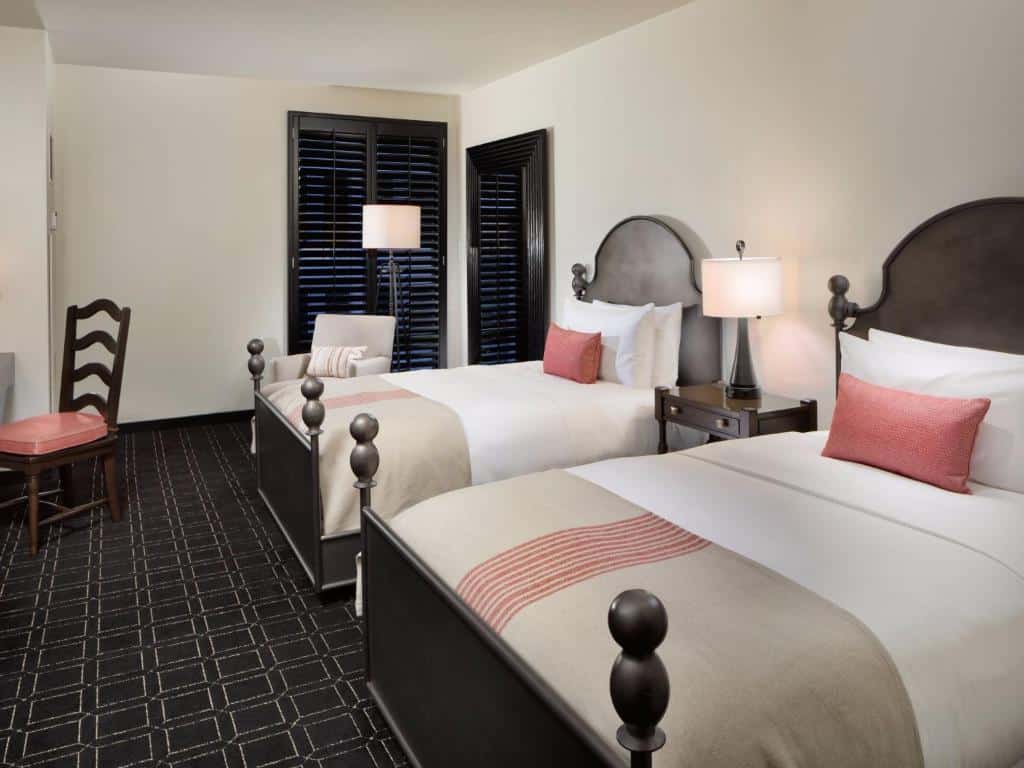 Quarto do Hotel Valencia Riverwalk com duas camas de solteiro nas cores marrom, branco e rosa, há uma porta para uma varanda, uma poltrona e uma cabeceira entre as camas com um abajur