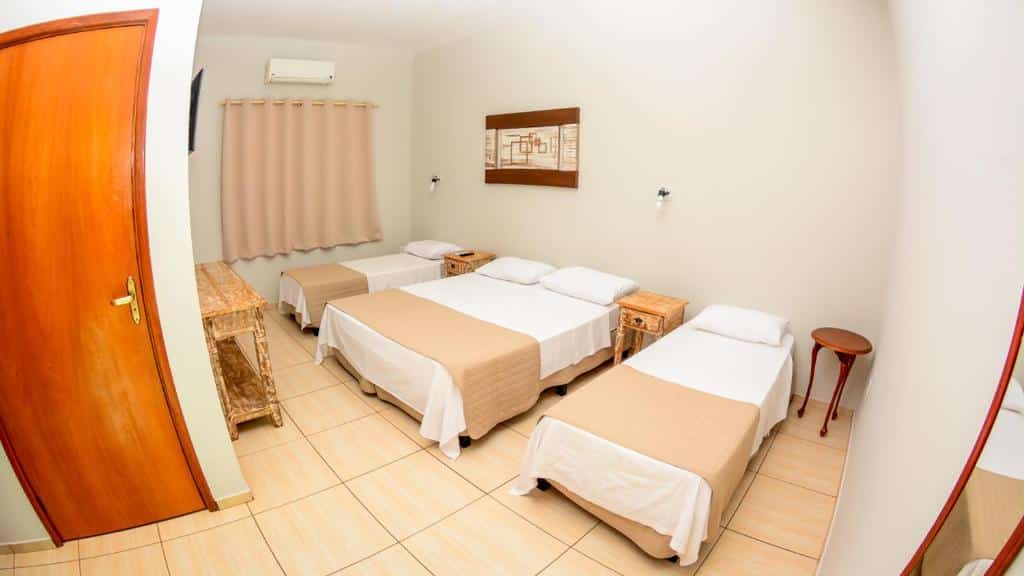 Quarto do hotel com 3 camas, uma de casal e duas de solteiro, com mesinhas do lado, quadro na parede branca, janela com cortina e uma porta de madeira.