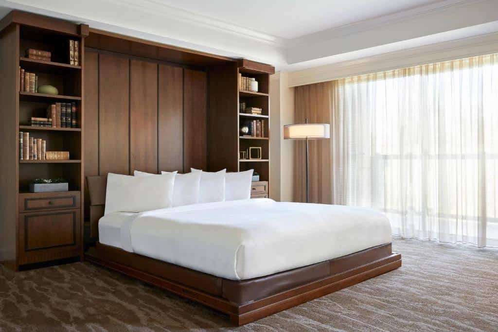 Quarto do JW Marriott San Antonio Hill Country Resort & Spa com uma cama de casal ampla, carpete marrom claro com detalhes em branco, uma sacada com cortinas, um abajur de chão e na cabeceira da cama há duas estantes com livros e itens de decoração