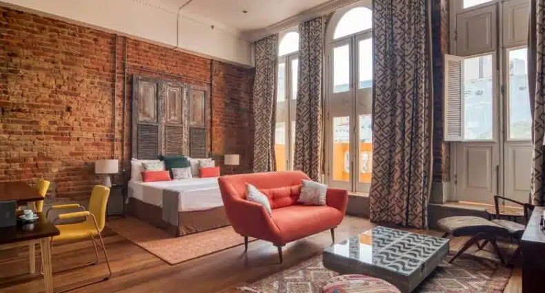 Um sofá na frente, com uma mesa de centro. Atrás uma cama de casal, no canto esquerdo uma mesa de escritório e no canto direito janelas com vista para a rua. Foto para ilustrar post sobre hotéis perto do Consulado Americano no Rio de Janeiro.