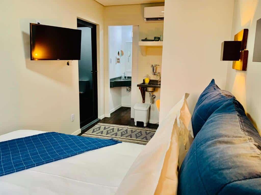 Quarto com uma cama com detalhes azul, uma tv na parede, uma pia ao fundo, tapete no chão e duas portas.