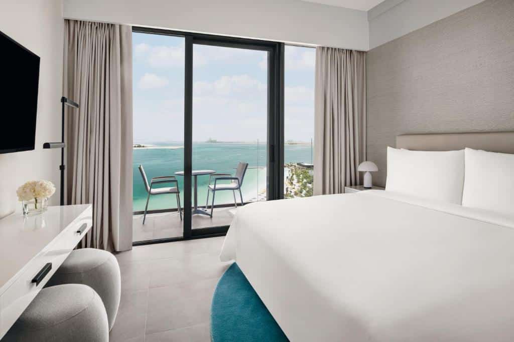 Quarto do Mövenpick Resort Al Marjan Island com uma varanda com vista para o mar, uma cama de casal com uma televisão de frente para a cama, com dois bufes cinzas e um tapete azul