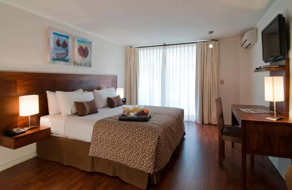 Quarto do Park Plaza Apart Hotel com cama de casal no centro, duas cômodas de madeira ao lado e uma mesa de madeira com cadeira em frente a cama. Representa hotéis baratos em Santiago.