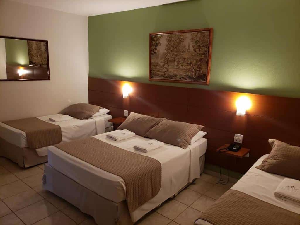 Quarto do hotel com três camas, uma de casal e duas de solteiro, com um quadro na parede verde e um espelho na parede branca.