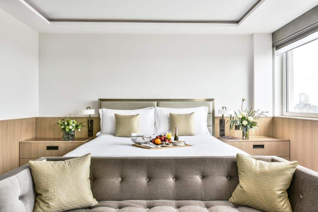 Quarto do Royal Lancaster London co uma janela com persiana, uma cama de casal com uma bandeja com frutas e bebidas sob ela, um sofá com dois lugares e duas mesinhas de cabeceira com vasos de flores