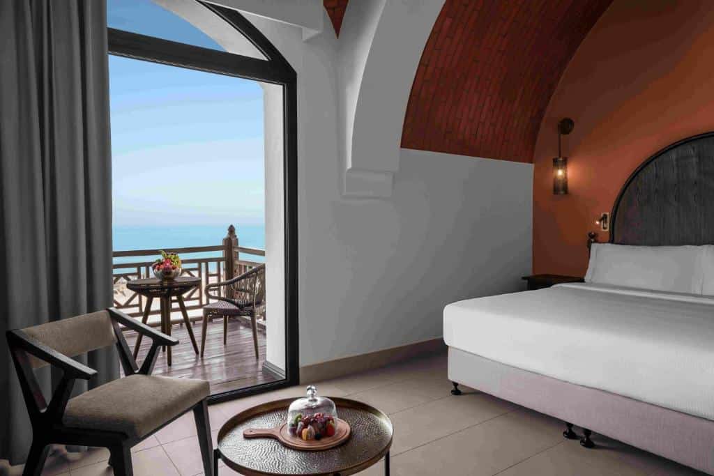 Quarto do The Cove Rotana Resort - Ras Al Khaimah com uma pequena varanda com vista para o mar, há uma cama de casal com uma cabeceira redonda, além de uma mesinha pequena com uma poltrona
