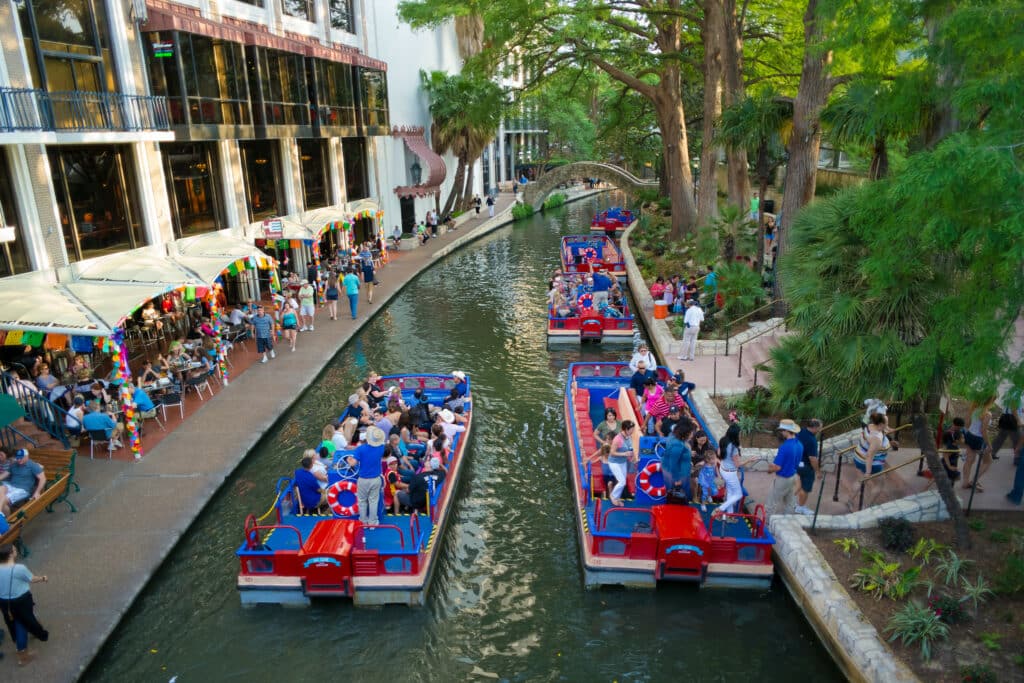 Um pequeno canal com embarcações em vermelho e azul, em uma das margens, há escadarias para as pessoas embarcarem e desembarcarem, do outro lado, há restaurantes e bares, todo o local é cercado por árvores