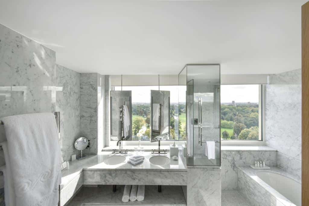 Banheiro do Royal Lancaster London com uma banheira com janelas amplas, uma pia com duas cubas, há toalhas brancas no local, para representar hotéis em Londres para brasileiros