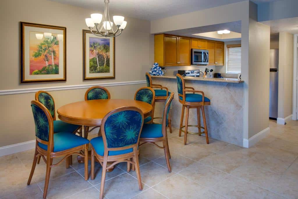 sala do FantasyWorld Resort com mesa e cadeiras de madeira com estofado azul com flores ao estilo praia e cozinha americana também com armários de madeira