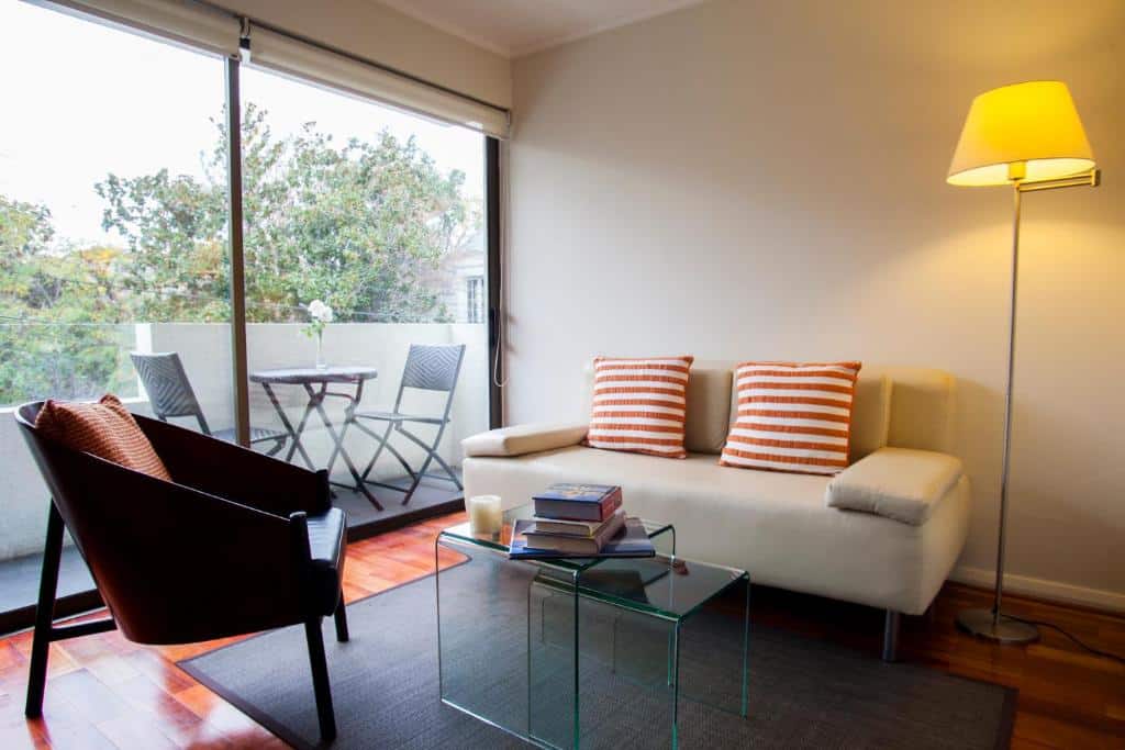 Sala de estar do Heidelberg Haus Apart Hotel com sofá branco a frente, mesa de vidro e uma cadeira. Representa aluguel de temporada em Santiago.