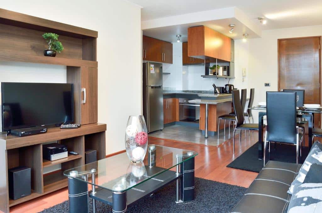 Sala do Norus Apartments Providencia com sofá, mesa de centro e TV em cima de uma estante e ao fundo uma mesa de vidro com cadeiras pretas e ao fundo uma cozinha. Representa aluguel de temporada em Santiago.