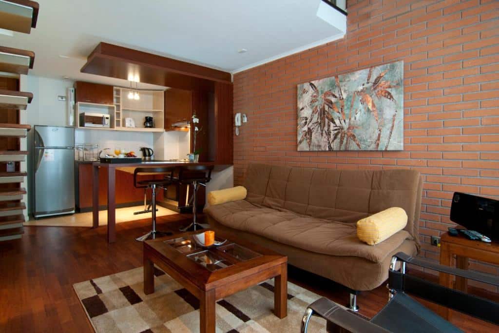 Sala de estar do Park Plaza Apart Hotel  com sofá marrom, uma mesa de centro de madeira e ao fundo uma cozinha. Representa aluguel de temporada em Santiago.