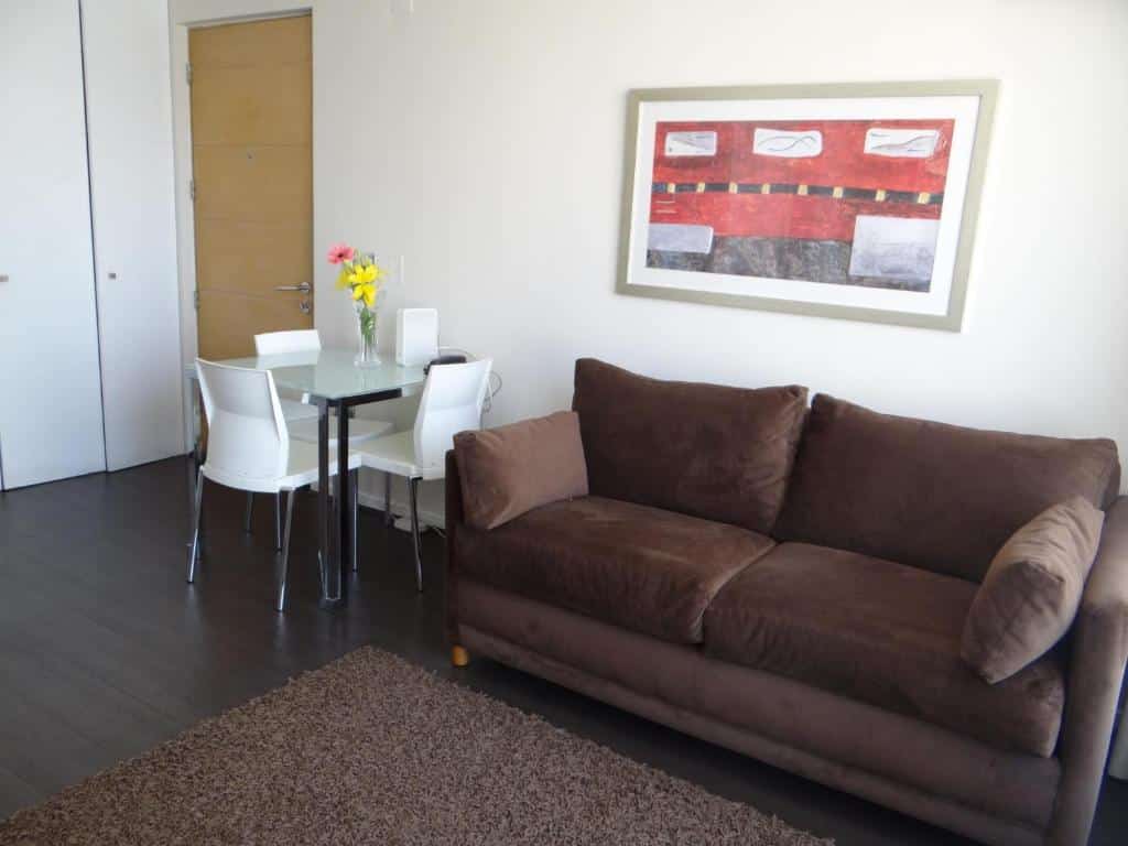 Sala do Rentasuite Lyon com sofá marrom do lado direito e uma mesa de vidro com quatro cadeiras brancas do lado esquerdo. Representa Airbnb em Santiago.
