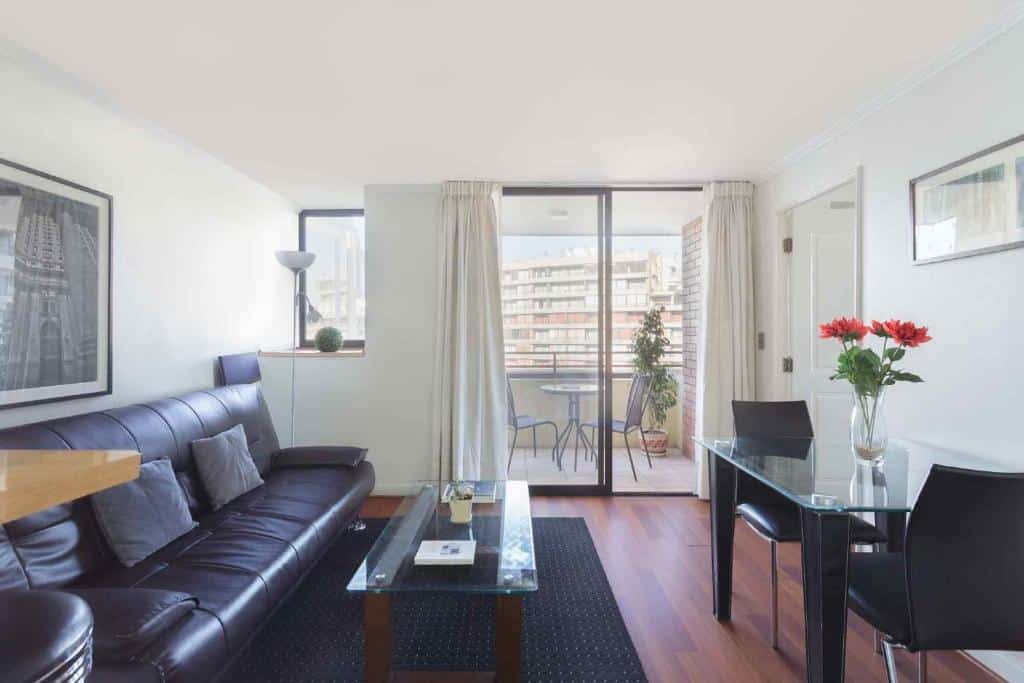 Sala do Santa Magdalena Apartments com sofá de couro preto do lado esquerdo, mesa de centro de vidro e uma mesa com duas cadeiras a frente. Representa Airbnb em Santiago.