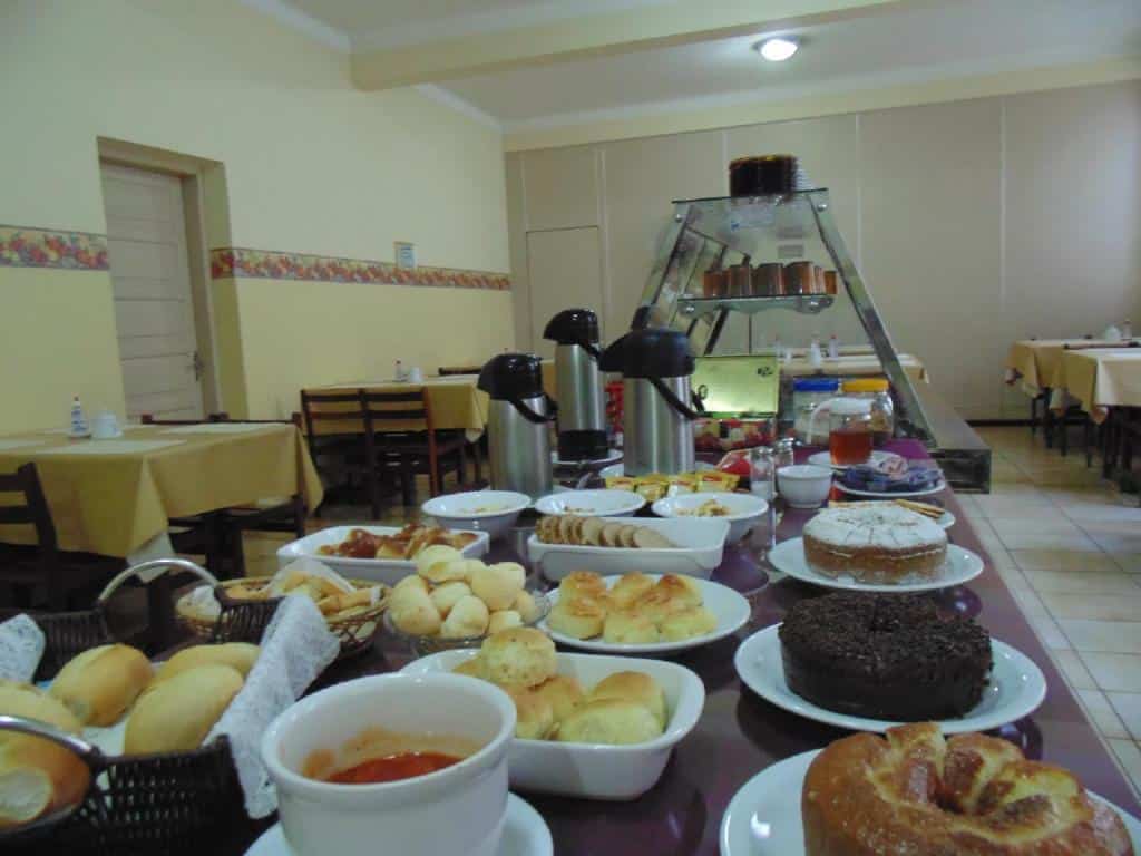 Mesa do café da manhã com várias opções de bolos, pães e café. Ao lado algumas mesas e cadeiras, ilustrando post Hotéis em Barretos.