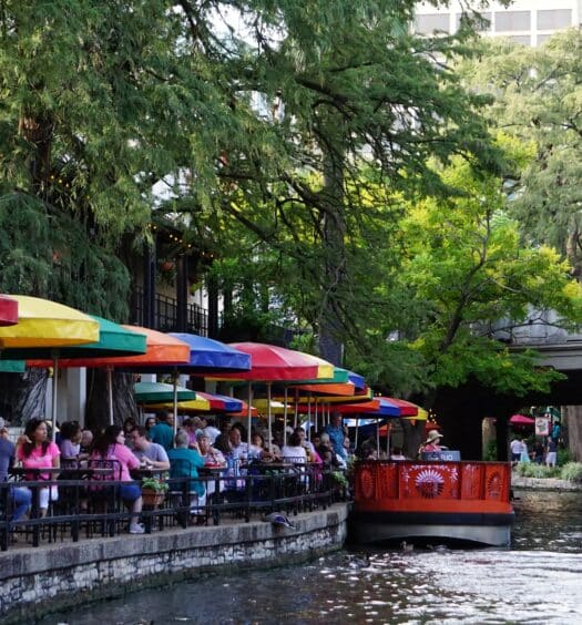 Um canal em San Antonio com algumas embarcações coloridas, há muitas árvores ao redor e uma ponte mais a frente, do lado esquerdo há restaurantes e pessoas sentadas às margens olhando a paisagem