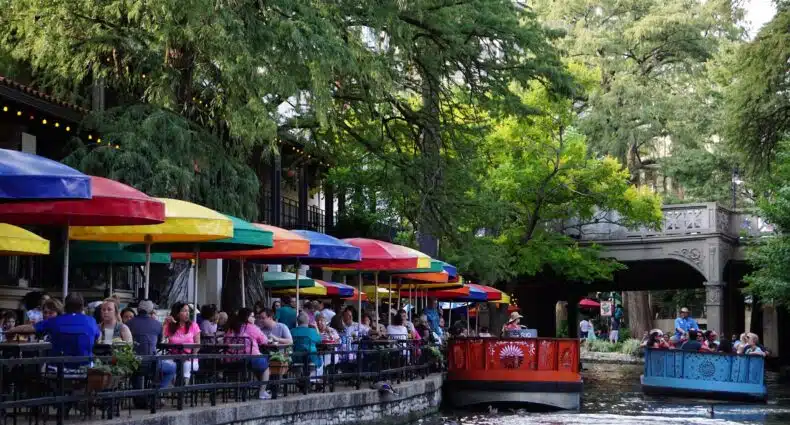 Um canal em San Antonio com algumas embarcações coloridas, há muitas árvores ao redor e uma ponte mais a frente, do lado esquerdo há restaurantes e pessoas sentadas às margens olhando a paisagem