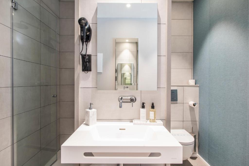 Banheiro do Sleep&Fly. A pia branca com espelho, secador de cabelo e amenidades de banho está no centro da foto. Do lado esquerdo fica o box com chuveiro, e do direito a privada.