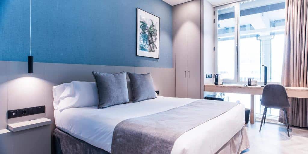 Quarto do Sleep&Fly, uma das recomendações de hotéis perto do aeroporto de Barcelona. A cama de casal tem mesinhas de cabeceira dos dois lados com luminárias. Ao lado da cama, um pequeno armário fica ao lado de uma mesa de trabalho com cadeira, cafeteira e uma janela com cortinas bege.
