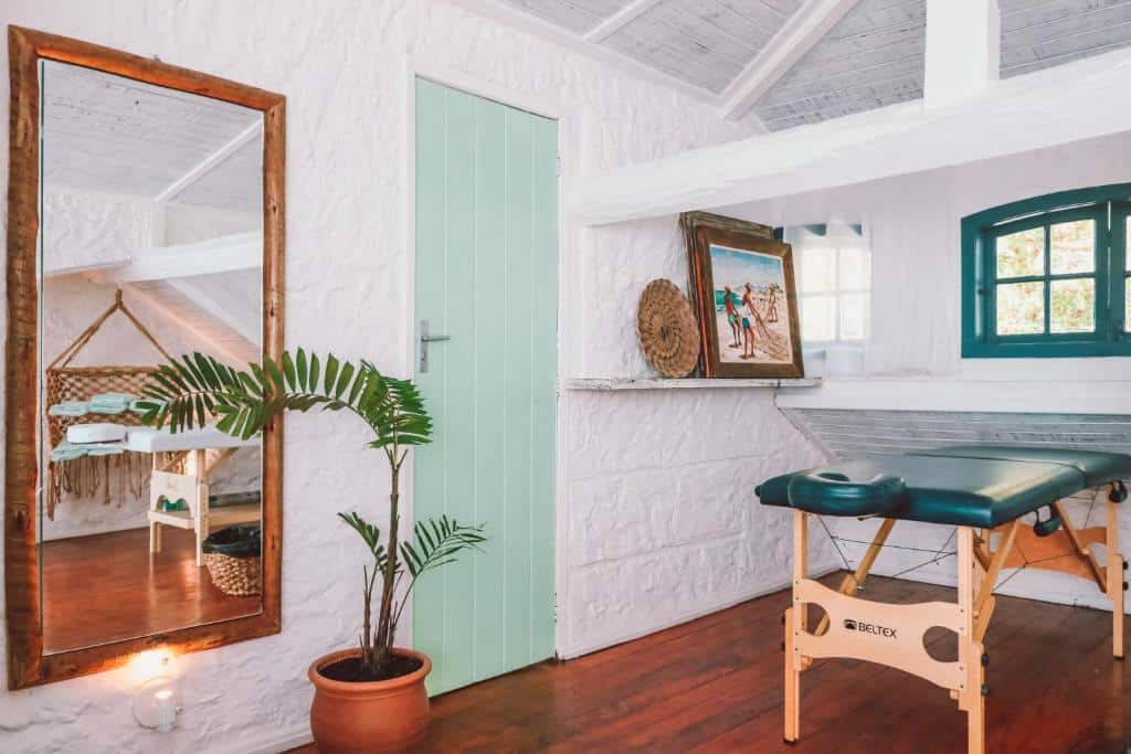 Sala de spa com paredes brancas, maca, porta e janelas pintadas em tons de verde, espelho, planta e quadro decorativo.
