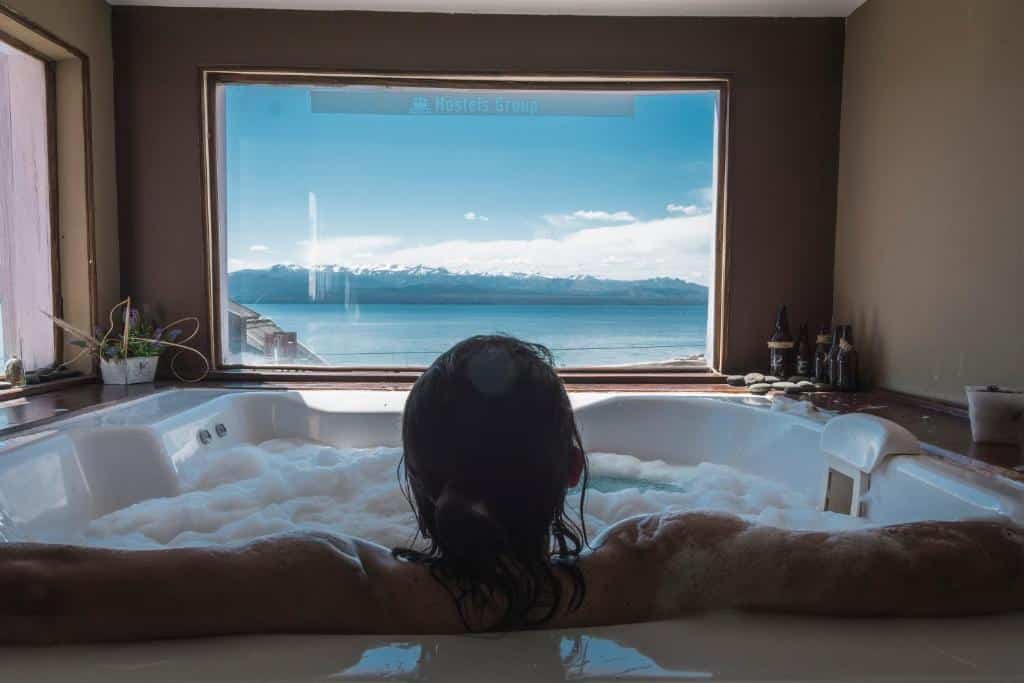 Mulher dentro de uma banheira de hidromassagem, com os braços abertos e apoiados na borda da hidro, de costas, olhando para uma janela de vidro com vista para um lago azulado e montanhas no horizonte. A hidro está com espumas e tem amenidades para banho nas bordas