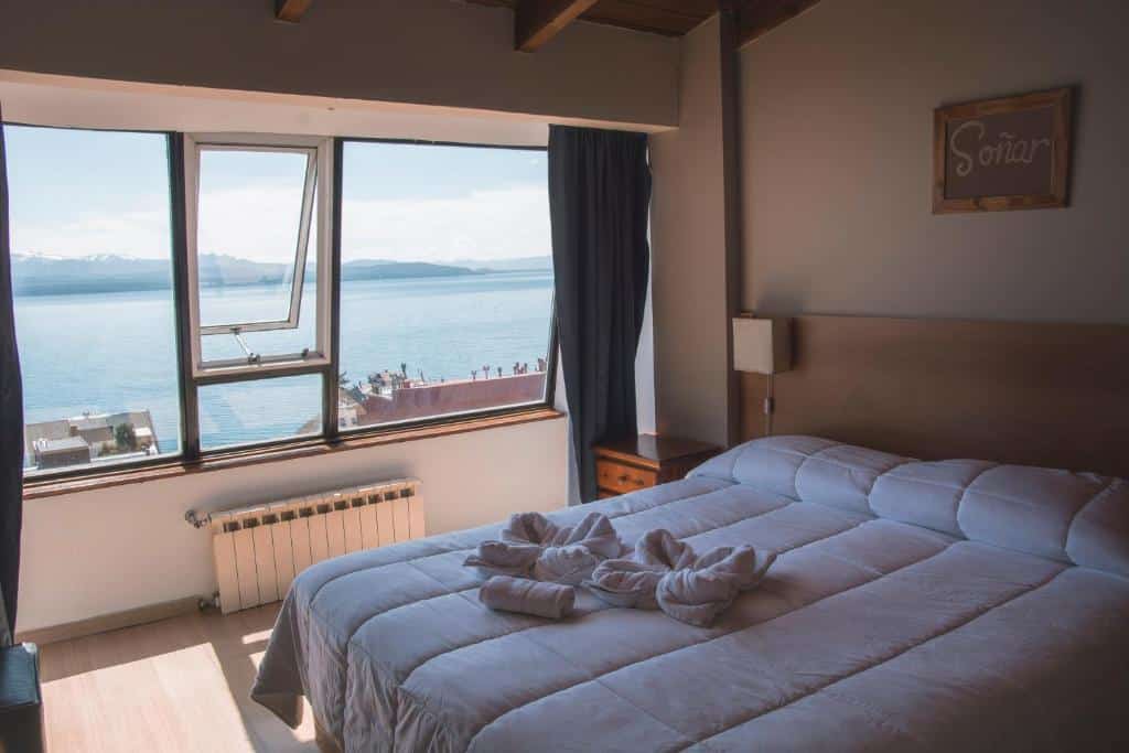 Quarto do Tangoinn, hostel onde ficar em Bariloche, com uma cama de casal com toalhas enroladas em cima e lençol branco e travesseiros e uma janela no lado esquerdo com vista para um lago cristalino e montanhas