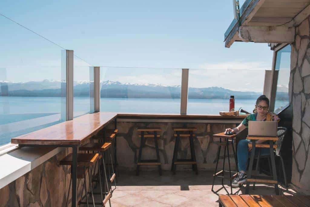 Área comum do Tangoinn Hostel Downtown, um dos lugares onde ficar em Bariloche, com um balcão com banquetas espalhadas, mesas do lado direito com uma mulher sentada mexendo no computador, e ao fundo vista para o lago