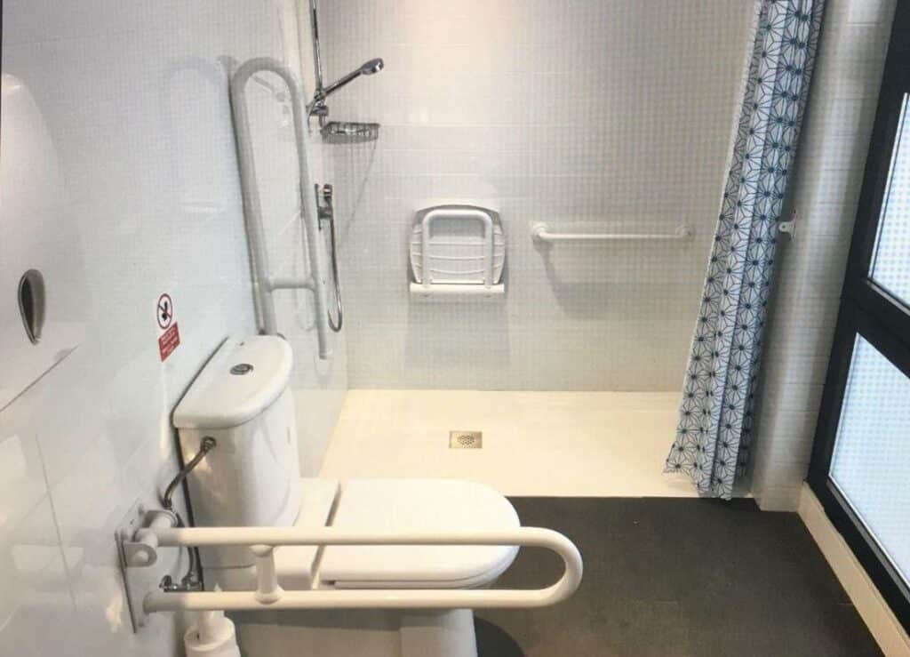 Banheiro do Ten To Go Hostel. A privada tem barras de apoio do lado, assim como a área de banho que tem cortina e assento de banho.