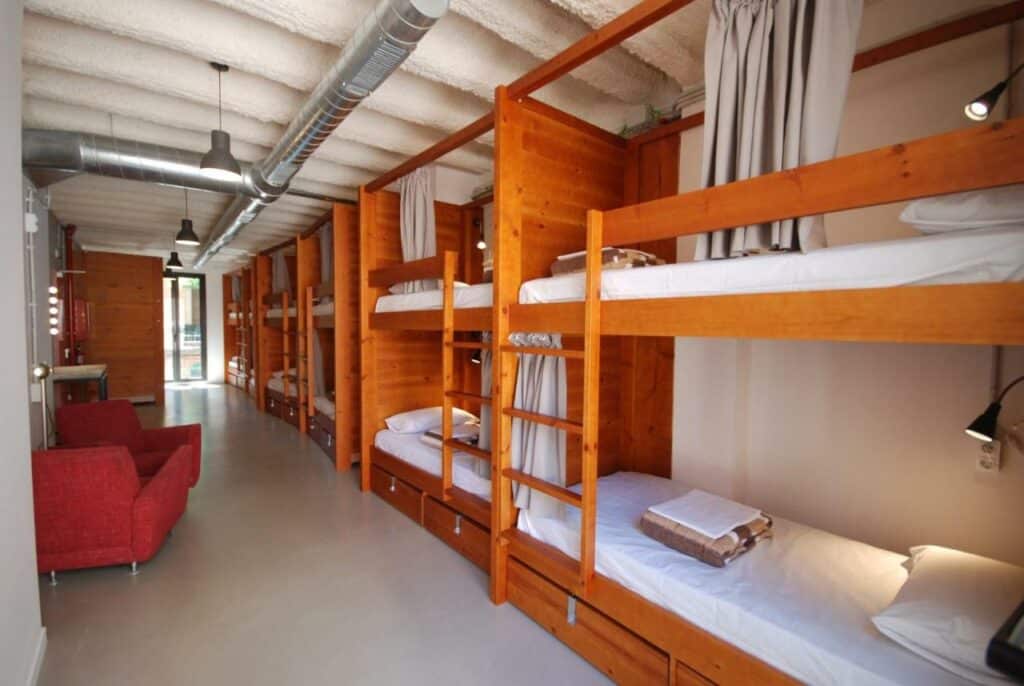Quarto do Ten To Go Hostel, uma das recomendações de hostels em Barcelona. Diversas beliches estão dispostas do lado direito do local com jogo de cama, toalhas e cortina de privacidade. Há uma poltrona vermelha do lado esquerdo, e ao fundo há uma porta de vidro.