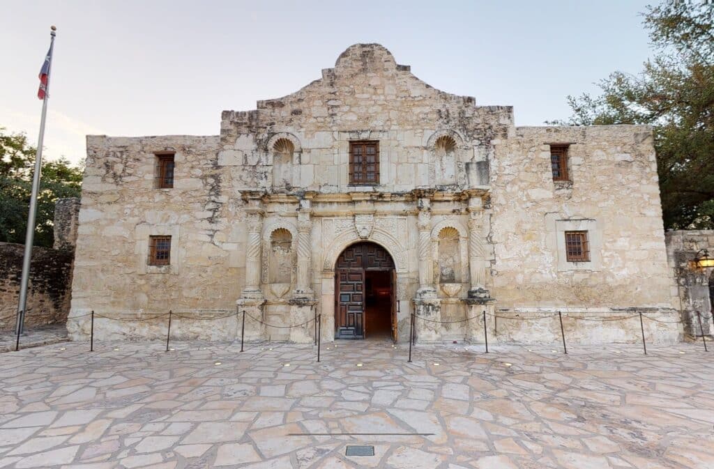 Um prédio antigo com quatro janelas, porta de madeira antiga, a construção é de um branco desgastado quase bege, há uma bandeira do Texas ao lado do local