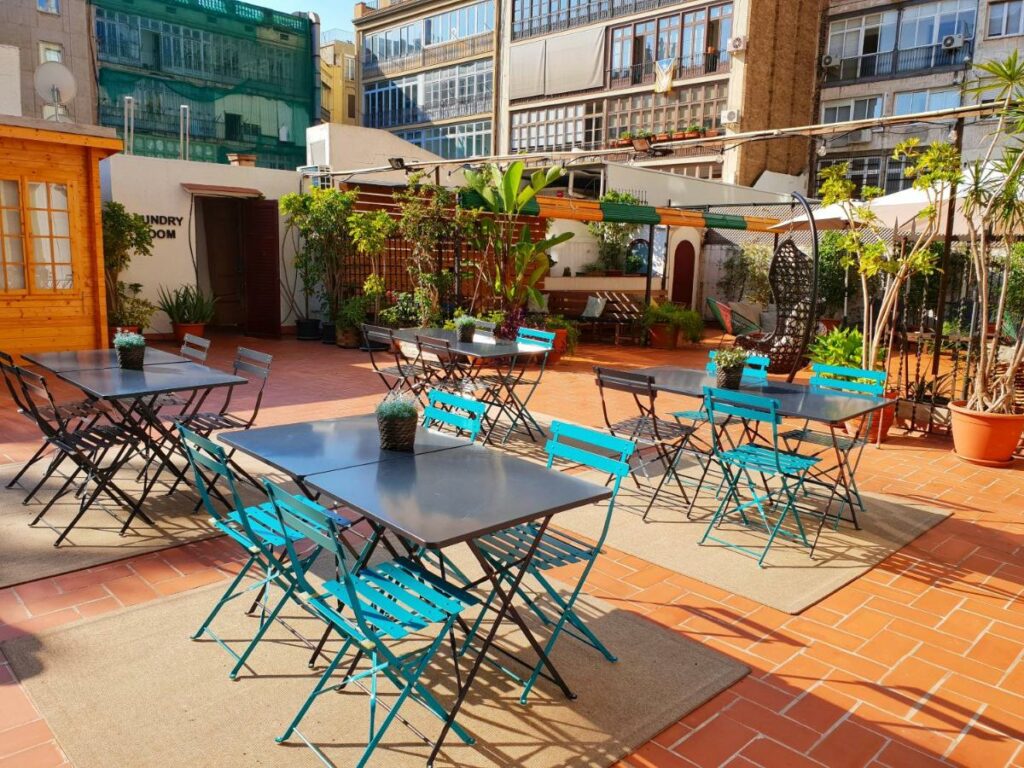 Terraço do The Central House Barcelona Gracia. Várias mesas com cadeiras estão dispostas pelo local, e ao fundo há plantas e mais lugares para sentar.